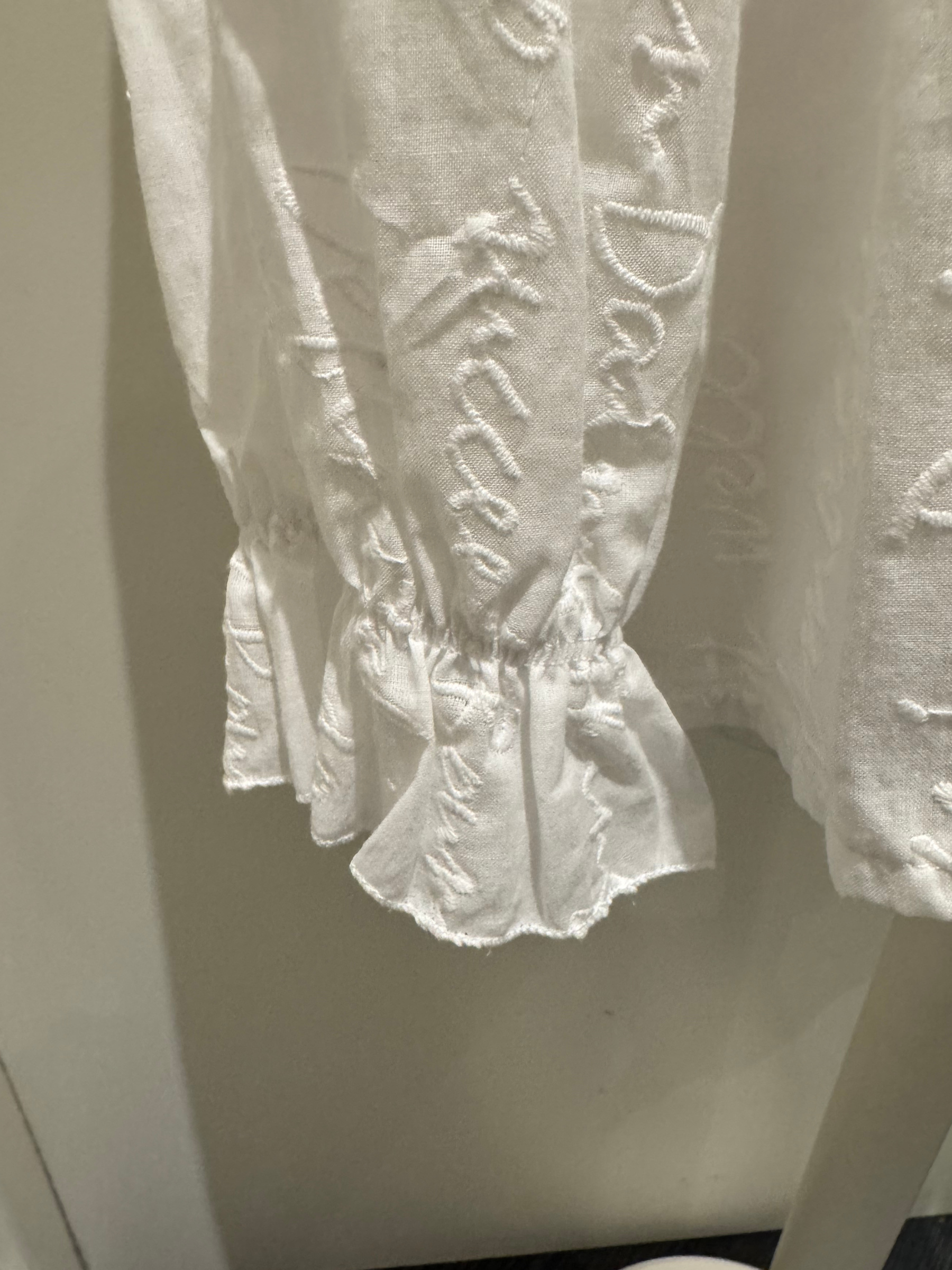 Deck by Decollage white embroidered cotton blouse featuring tonal script text, a split neckline, and ruffle cuffs. A breathable and elegant top ideal for smart-casual dressing.