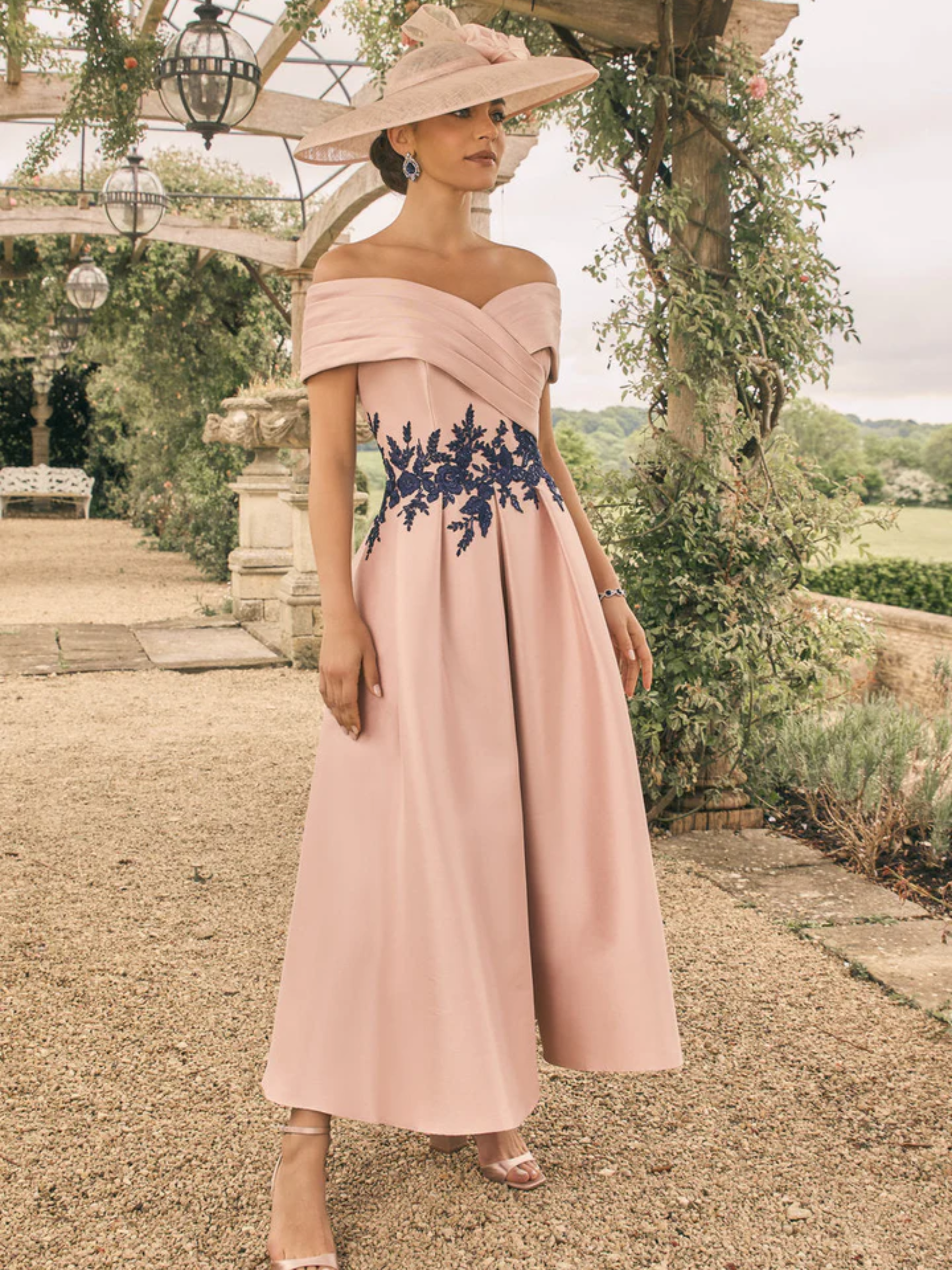Veni Infantino 992709B Blush/Navy Dress with an off-shoulder crossover neckline, navy floral lace applique detailing and an A-line structured midi skirt - an elegant blush occasion dress perfect for weddings and special events.