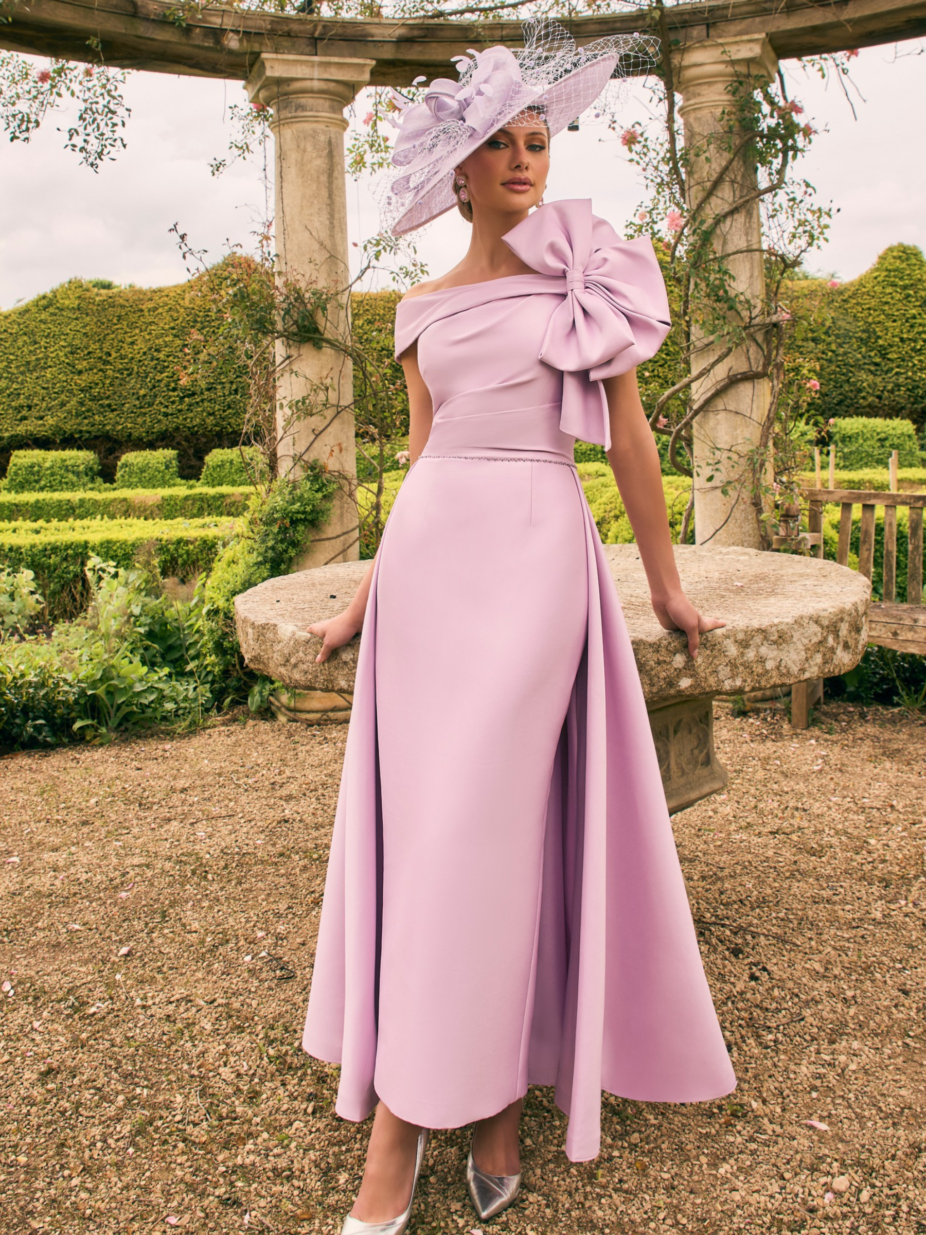 Veni Infantino 992731 Lilac crepe dress with off-the-shoulder neckline, oversized bow and flared A-line skirt. Elegant occasion dress for weddings and special events.